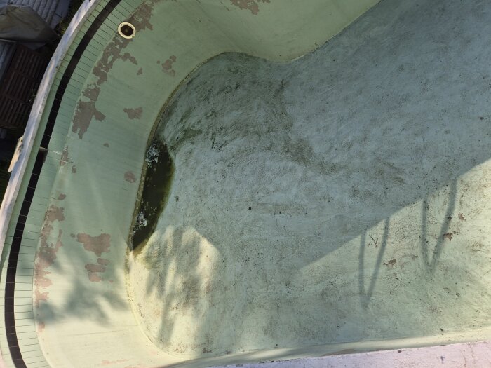 Sliten, gammal betongpool med flagnande färg och lossnande kakelplattor. Vatten och fuktfläckar syns på botten av poolen.