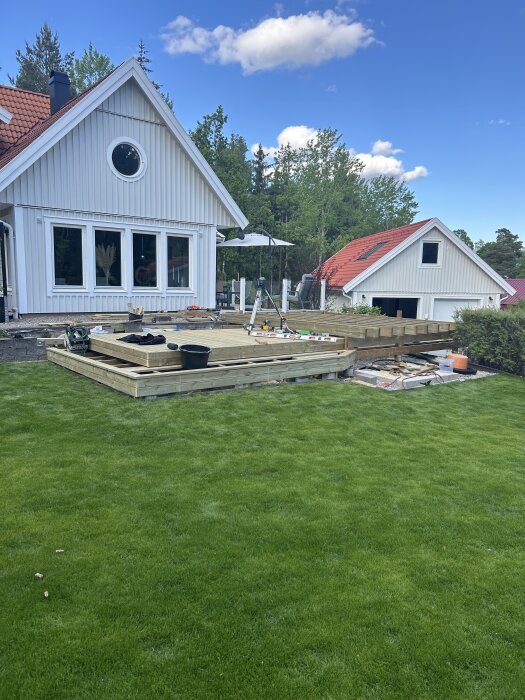 Altan under byggnation vid ett vitt hus, med verktyg och material utspridda på den halvfärdiga strukturen, omgiven av grön gräsmatta.