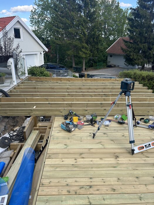 Altanbygge med verktyg och träplankor, laser på stativ, omgiven av hus och träd i bakgrunden.