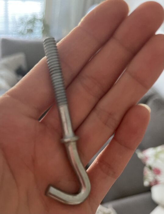 Hand håller en metallkrok med gängor, används för att hänga upp föremål som en kristallkrona i taket.