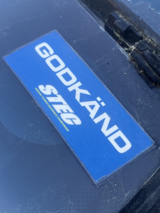 Blå dekal på en bil med texten "GODKÄND" och "STEC" i vitt, placerad på en dammig vindruta.