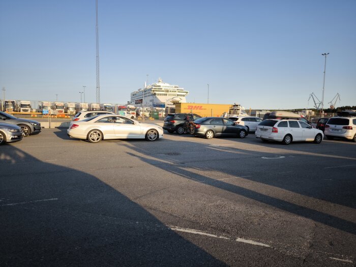 Bilar parkerade nära en färja vid kajen, med blå himmel och solnedgång som kastar långa skuggor över parkeringsplatsen.