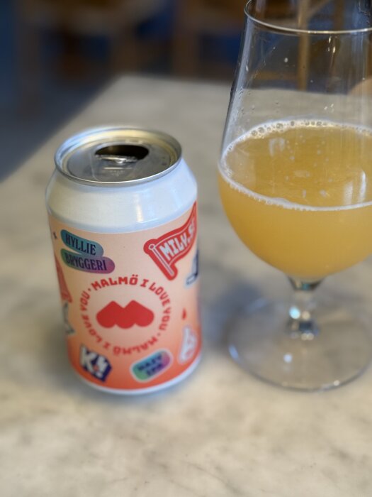 Ölburk med texten "Hyltie bryggeri, Malmö I love you" och ett glas med ljusgul öl på ett marmorbord.