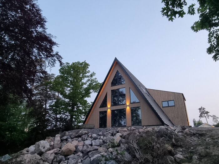 Triangulärt hus i trä omgiven av träd, placerat på en stenig sluttning med en klar himmel i bakgrunden.
