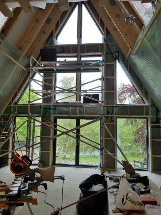 Renoveringsprojekt med ställning inomhus, stora fönster mot trädgård. Verktyg och material synliga i förgrunden.