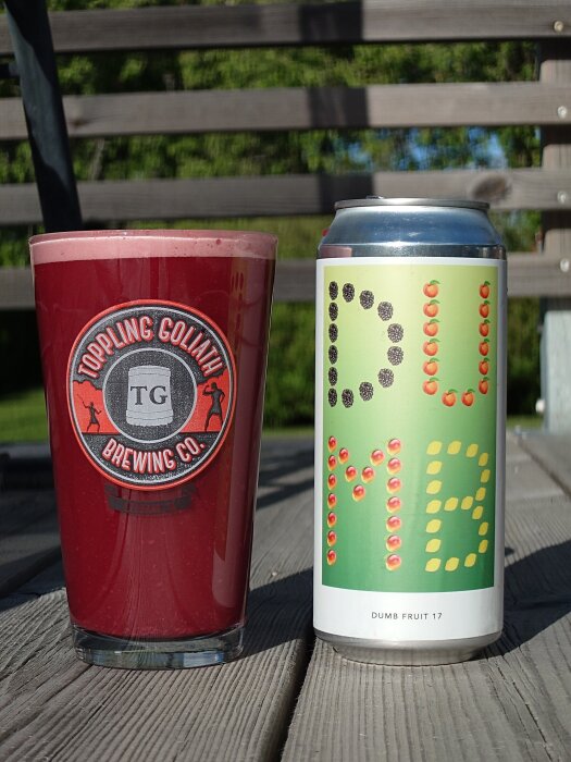 Glas med röd suröl bredvid en ölburk med etiketten "Dumb Fruit 17" på en träyta utomhus.