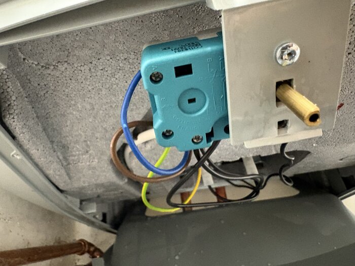 Elektrisk komponent med blå plastkåpa och flera anslutna kablar, monterad inuti en maskin. En guldig axel sticker ut från en metallfäste.