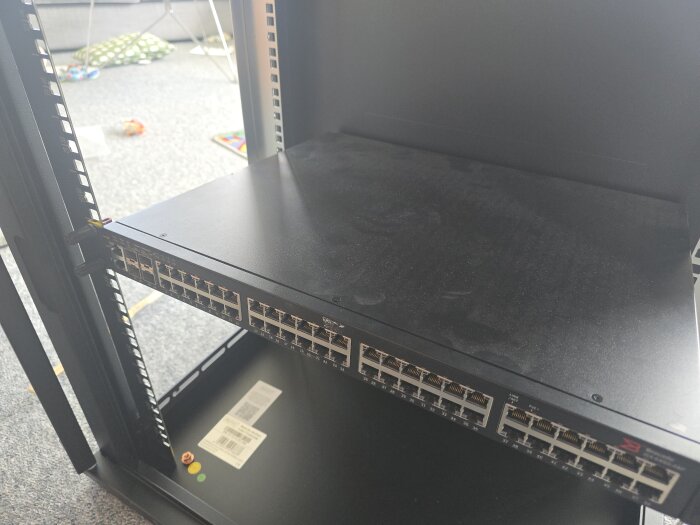 Nätverksswitch monterad i ett svart serverrack, synliga portar framtill. Bakgrunden visar en del av ett rum med leksaker på golvet.