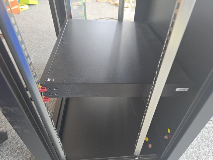 Svart serverrack i ett rum. Två horisontella hyllor syns, med några små monteringsdetaljer på sidorna. Grått golv och färgglada objekt i bakgrunden.