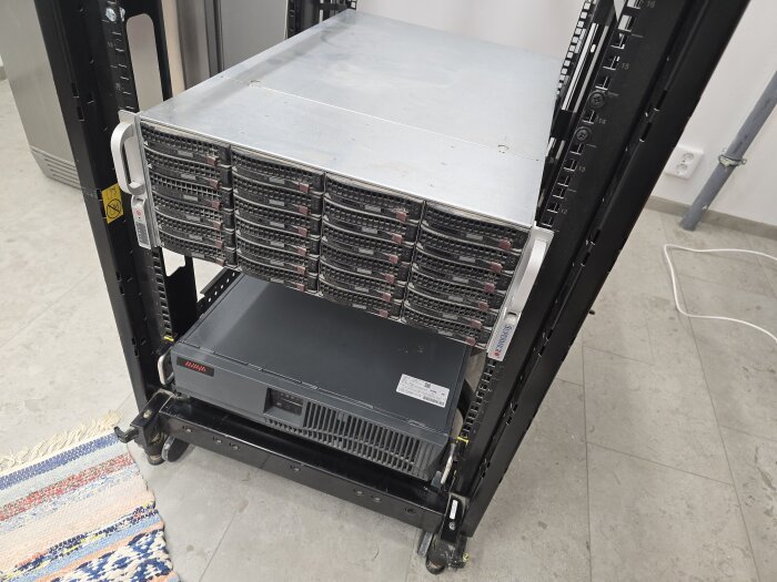 Serverrack i maskinrum med 4U-chassi ovanpå UPS-enhet, omgiven av golvmatta och vägguttag i bakgrunden.