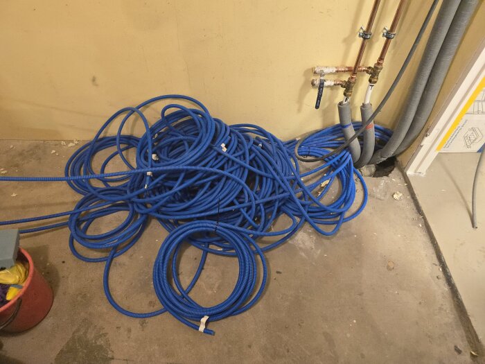 Blå flexslangar med nätverkskablar ligger huller om buller på ett betonggolv nära vattenrör i ett hobbyrum.