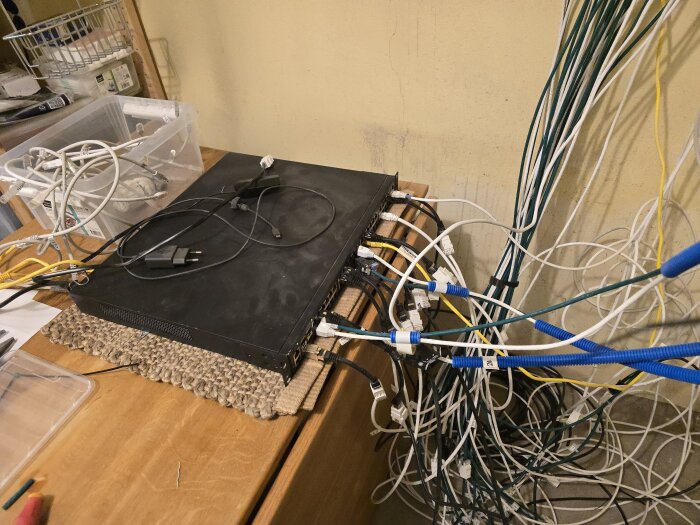 En server med många kablar och sladdar anslutna, placerad på ett bord i ett hobbyrum fyllt med nätverksutrustning och förvaringslådor.