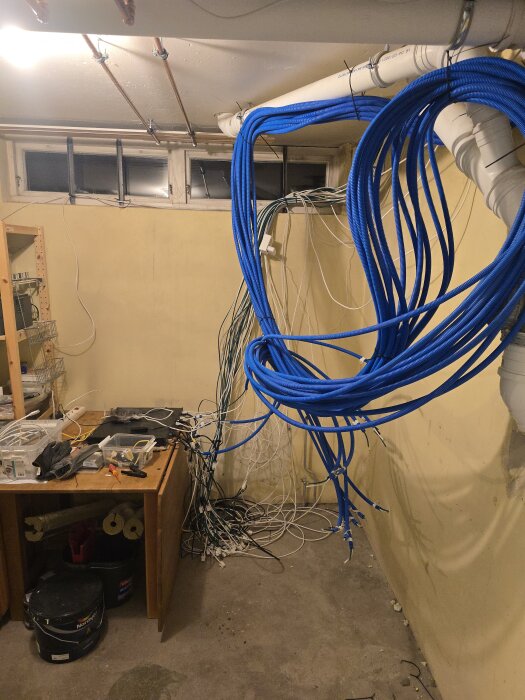 Blå kablar och rör i hobbyrum; nätverksinstallation under arbete med synliga kablar och verktyg på bordet, fläktar och fönster i bakgrunden.