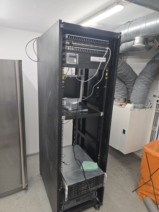 Serverrack med inkopplade kablar och elektronisk utrustning, omgiven av ventilation och vit vägg.