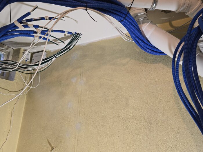 Blå flexslangar och kablar fästa i taket i ett rum med gulaktig vägg. Ventilationsrör syns ovanför. Taken verkar nyligen ha målats.