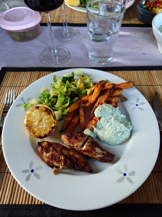 Grillad kycklingfilé, sötpotatispommes, grillad citron, sallad och basilikasås på en tallrik.