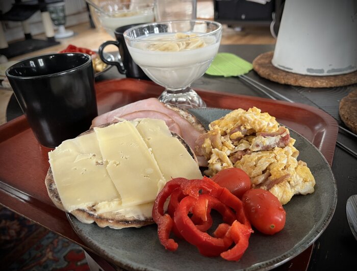 Söndagsfrukost på bricka med kaffe, skinkmacka med ost, äggröra med bacon, tomater, paprika och yoghurt med banan i skål.