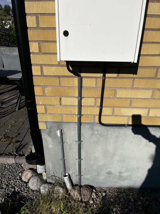 Elskåp monterat på en gul tegelvägg med kabelskydd som går ned mot marken, omgiven av stenar och grönska.