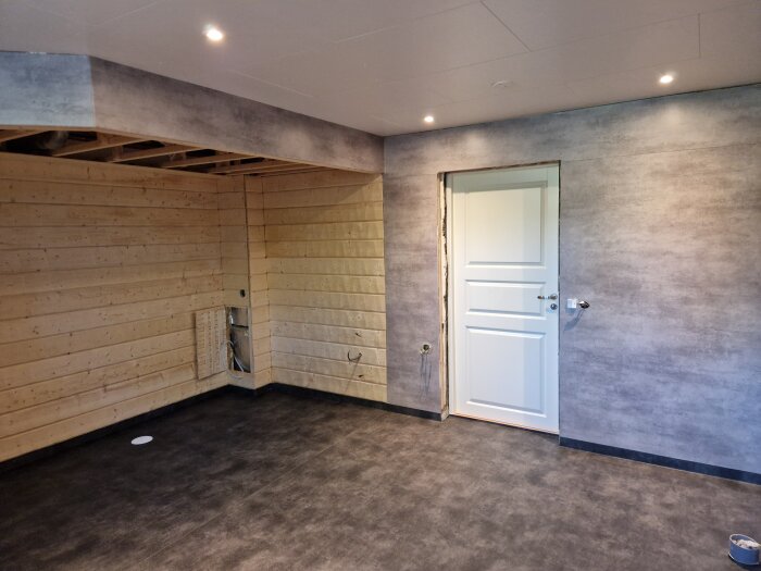 Badrum med mörka golv och grå väggar, en vit dörr och träpaneler under renovering.