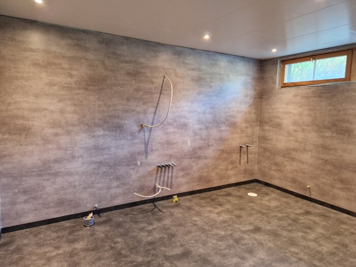 Badrum med mörka väggar och golv, installationsrör synliga, fönster överst på väggen, förberedelser för renovering.
