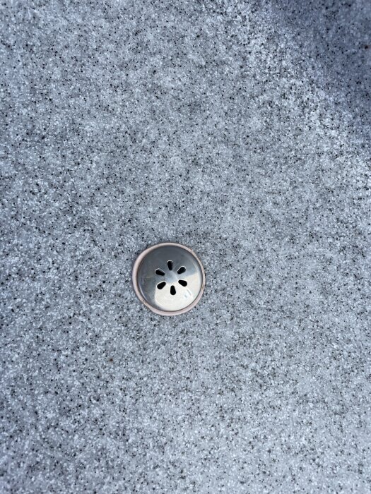 Metallplatta med sexslitsade hål monterad i grå, knottrig yta, kanske för luftbubblor i spa eller pool.