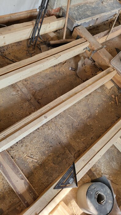 Byggrenovering med synliga träbjälkar, golvspån, en vinkelhake och rörliga byggmaterialverktyg på en dammig arbetsyta.