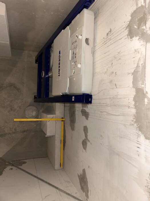 Bild av Geberit fixtur installerad på väggen, med ett måttband som mäter avståndet till avloppsröret och väggen. Väggar redo för renovering.