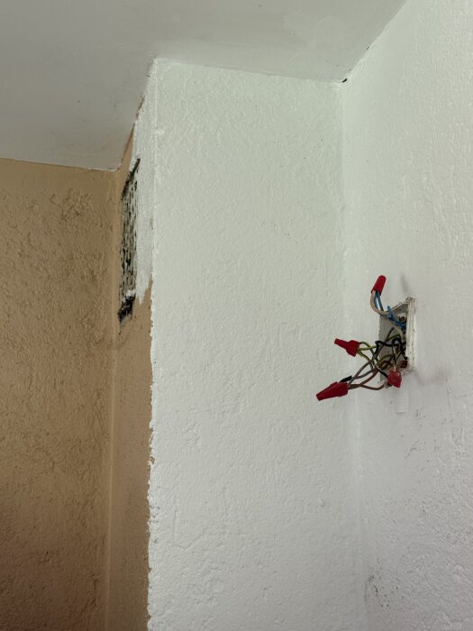 Övergång mellan två väggar i ett kök med synliga elkablar och en ventilation i hörnet av den ljusa väggen.