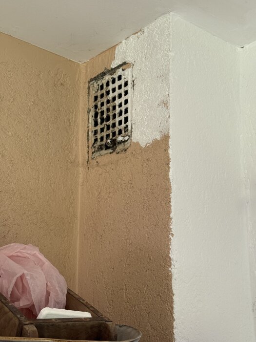 Ventil på väggen med blandad vit och brun färg, omgiven av skrovlig yta. Två hörn synliga, samt en korg med rosa plastpåse under.