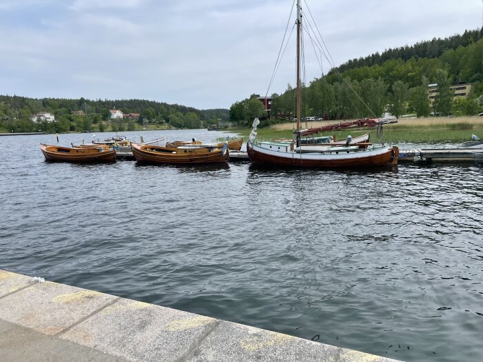 Flera träbåtar ligger förtöjda vid en brygga vid en skogig kustlinje under en molnig himmel.