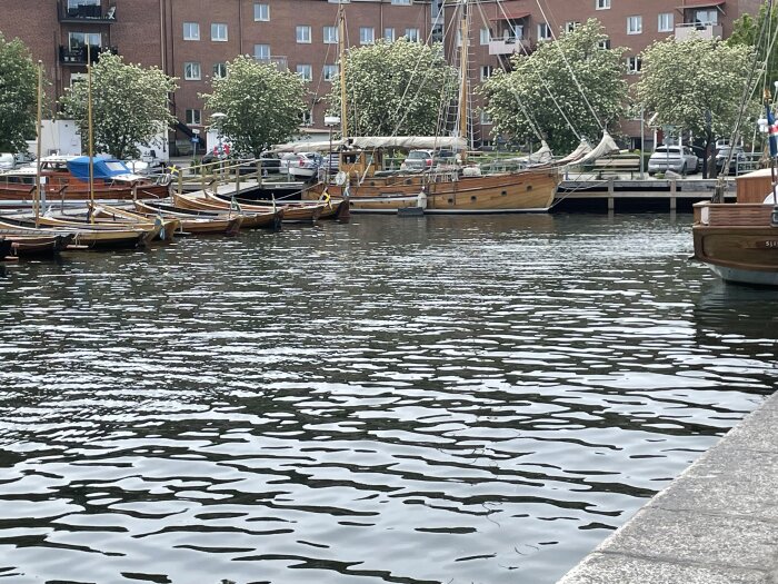 Båtar ligger förtöjda vid en brygga i en hamn, med byggnader och träd i bakgrunden och vatten som glittrar i förgrunden.