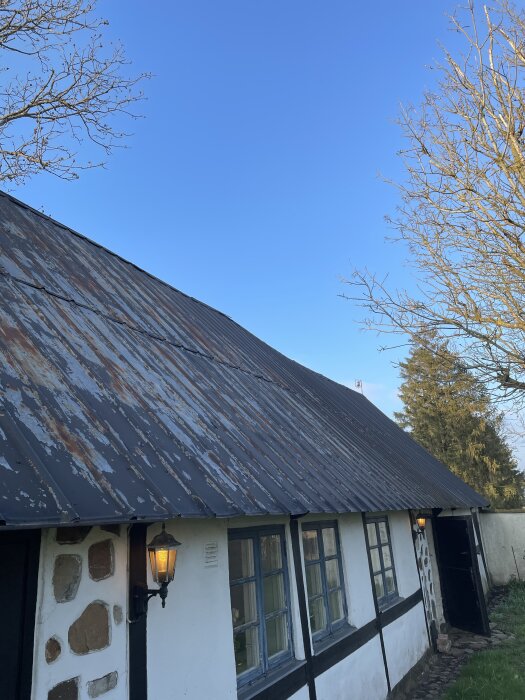 Gammal skånelänga med vitputsade väggar, svart plåttak och lampor på fasaden, omgiven av trädgrenar och klar blå himmel.