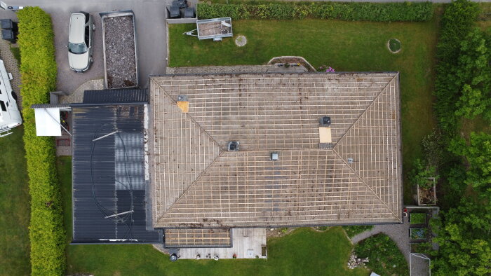 Luftfoto av ett hus där takpannor är borttagna, visar takstolar och en avställt container med rivningsmaterial på en grusuppfart.