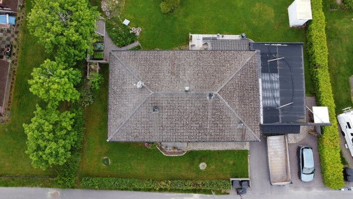 Flygbild av ett hus med brun takpannor och grön trädgård. En lastbil och en bil är parkerade på uppfarten bredvid huset.