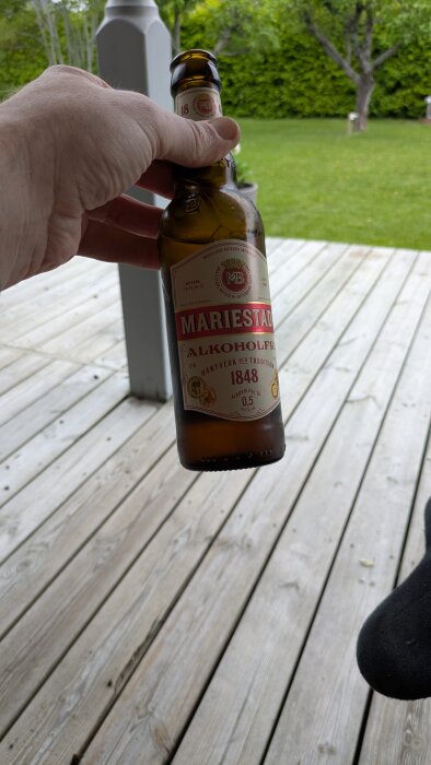 En person håller i en flaska Mariestad alkoholfri öl, stående på en altan med gräsmatta i bakgrunden.