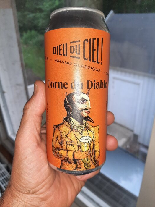 En hand håller en ölburk med etikett från Dieu du Ciel! Corne du Diable, med en illustration av en djävul som håller ett ölglas.