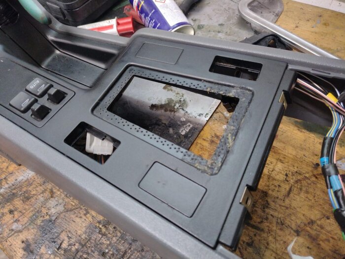 Centerkonsol från Volvo 850 med synliga sprickor och skador, borttagen från bilen och placerad på en arbetsbänk med verktyg och kablar.