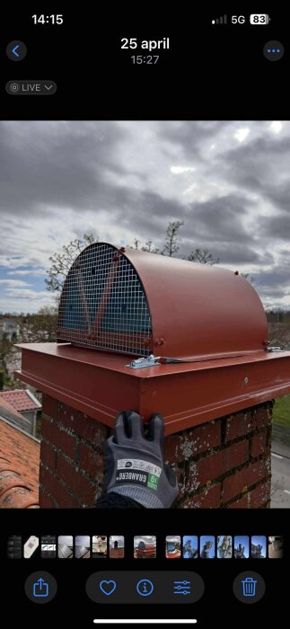 Murad skorsten med rött gallerlock och person i arbetshandske som justerar lock över öppning under molnig himmel.