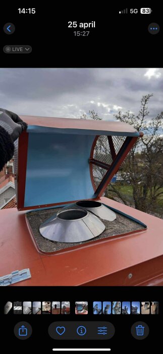 Röd ventilationskåpa med öppet lock och dubbla ventiler på ett tak, omgiven av träd och molnig himmel.