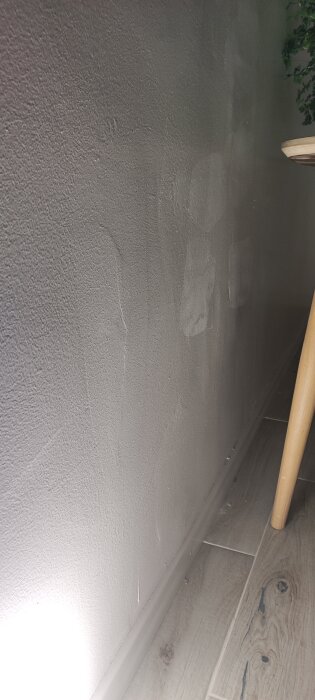 Ojämnt putsad vägg i källare belyst av ficklampa, visar textur och spår av spackling. Träbord synligt i hörnet, ligger på klinkergolv.