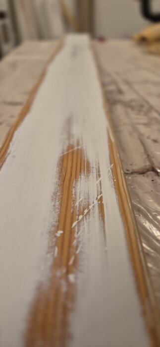 Närbild av ett slipat trappräcke med synliga lager av vit färg och trä, fotograferat i en inomhusverkstadsmiljö.