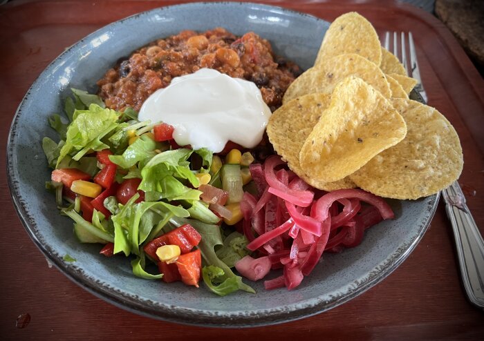 En tallrik chili con carne med grekisk yoghurt, sallad, picklad rödlök och nachos.