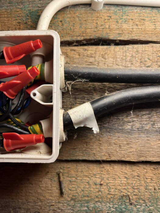 Elektrisk kopplingsdosa med röda wire nuts på svarta och jordade ledningar, monterad på trävägg i äldre byggnad.
