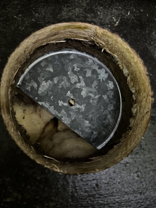 Rund öppning i en vägg, med ett lager av cirka 1 mm plåt bakom asfaboard, ovanför ett fönster.