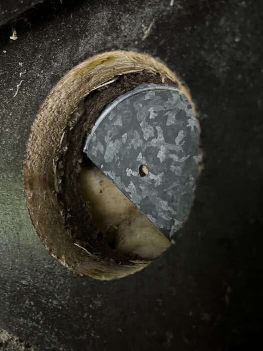 En cirkulär öppning i ett väggmaterial visar en ca 1 mm tjock plåt bakom asfafiberboard, ovanför ett fönster.