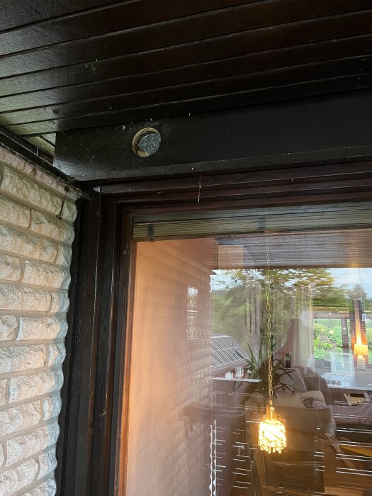 Bilden visar en del av en fasad med träpanel och tegel över ett fönster. Ett plåtföremål syns ovanför fönstret, omgiven av träpanelens skarvar.
