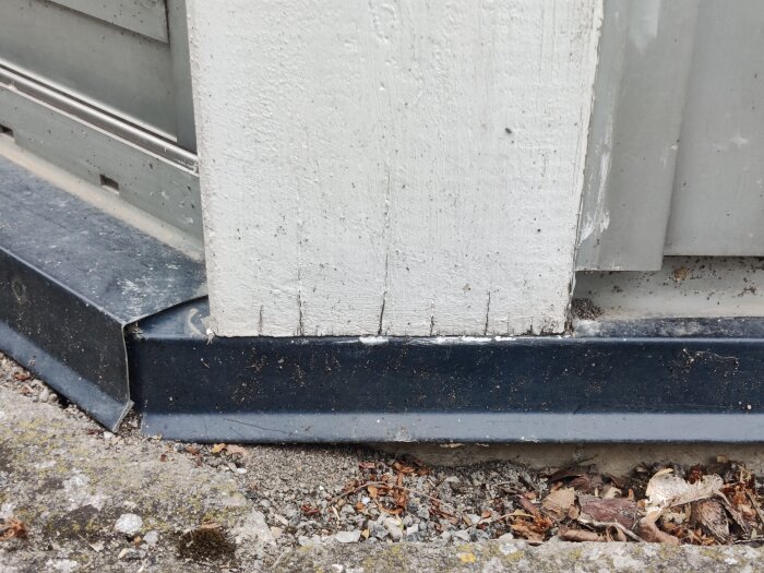 Murken limträstolpe stående på en plåt vid grunden av ett uterum, med tecken på vattenupptagning och omgivande grus och löv.