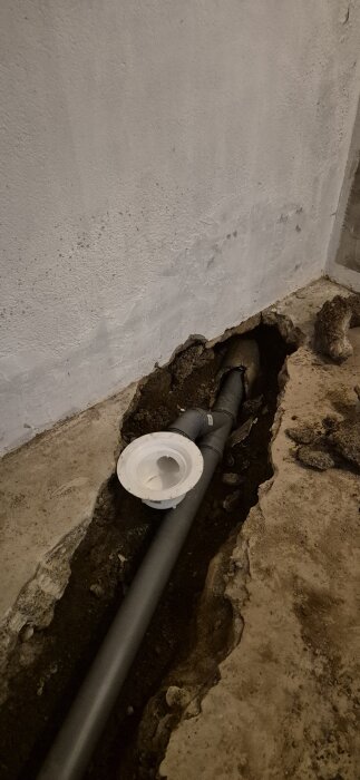 Grå avloppsrör installerat i en uppgrävd ränna i betonggolv, med vita rördelar synliga. Relaterat till köksstamsbyte.