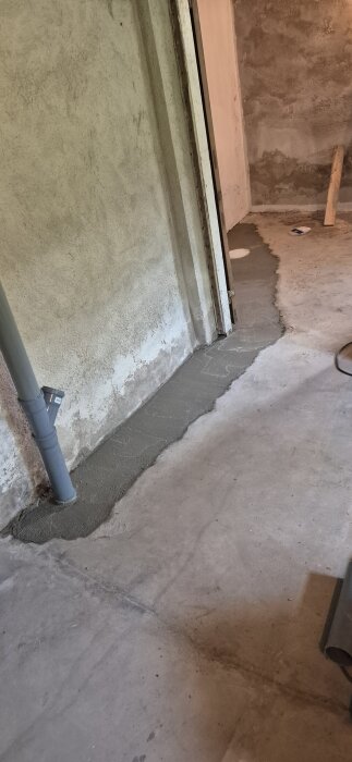 Ny cementlagd köksstam, nyläggning i ett renoveringsprojekt. Grå avloppsrör synligt längs väggen mot en cementgolv kanal.