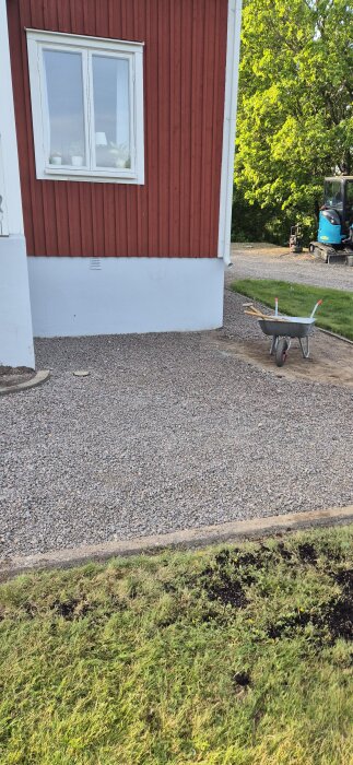 Röd trähusvägg med vitt fönster, grusbelagd mark och skottkärra med verktyg. Grävmaskin i bakgrunden.